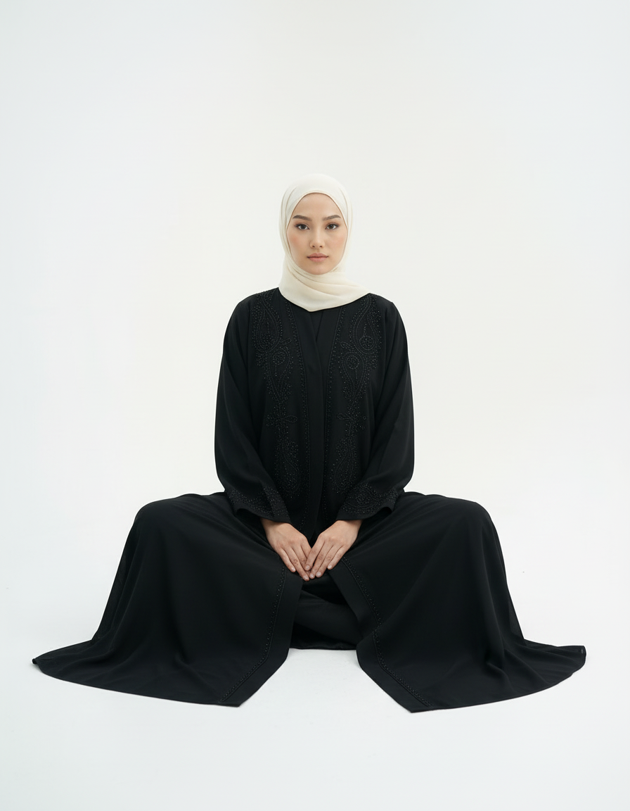 Seated floor pose showing wide drape and hem spread, black abaya and cream hijab, studio white background.